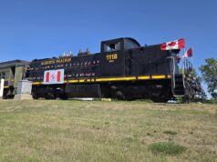 2026 Kin Canada ‘Ride the Rails’ Convention Held in Stettler Black steam locomotive with Canadian flags on the cabin, Alberta Prairie 1118, on a grassy rail yard under a clear blue sky.