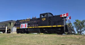 2026 Kin Canada ‘Ride the Rails’ Convention Held in Stettler Black steam locomotive with Canadian flags on the cabin, Alberta Prairie 1118, on a grassy rail yard under a clear blue sky.