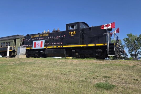 2026 Kin Canada ‘Ride the Rails’ Convention Held in Stettler Black steam locomotive with Canadian flags on the cabin, Alberta Prairie 1118, on a grassy rail yard under a clear blue sky.