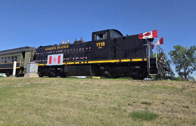 709035653_1484000333766776_7677285400581360884_n Black steam locomotive with Canadian flags on the cabin, Alberta Prairie 1118, on a grassy rail yard under a clear blue sky.