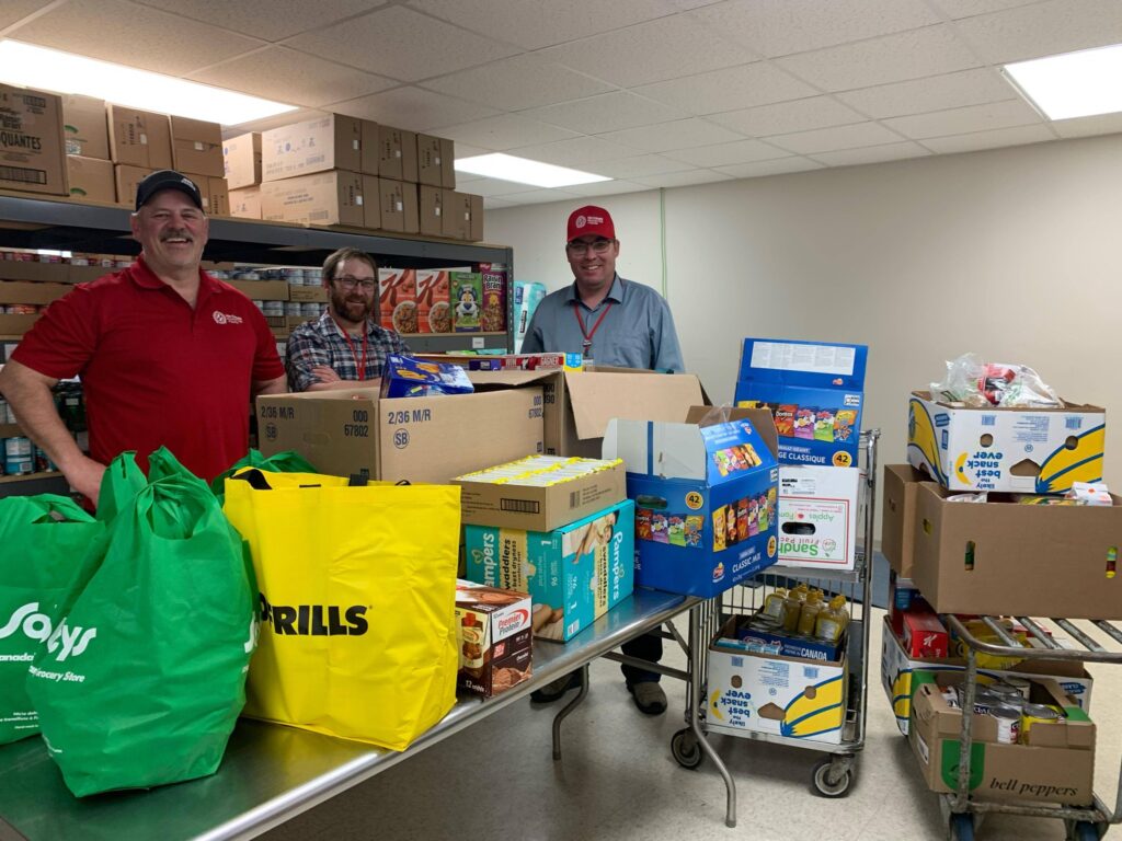 Three volunteers in a cluttered donation room, smiling at the camera among stacked boxes and grocery bags in the foreground.