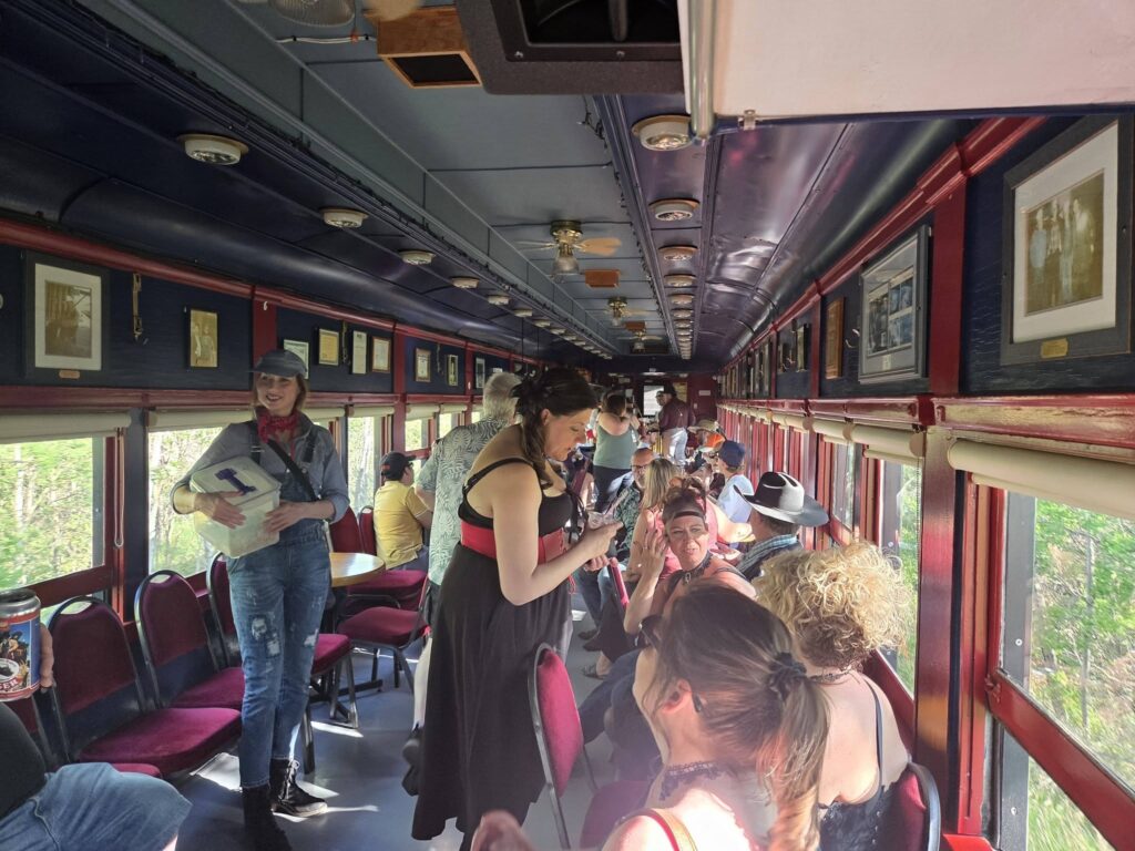 Crowded vintage train carriage with burgundy seats, passengers standing and chatting, framed photos along dark blue walls.