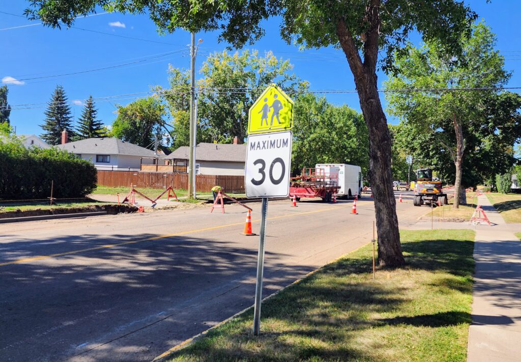Residential street under construction with orange cones, signs, and construction equipment; 30 mph speed limit posted under a pedestrian crossing sign.
