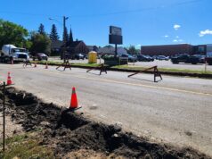 Stettler Back-to-School Traffic: 50 Ave Construction & Detour Map Construction work on a city street with orange cones and red barricades; a deep trench runs along the road and a large cement mixer truck is parked nearby, with buildings and trees in the background.