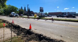 Stettler Back-to-School Traffic: 50 Ave Construction & Detour Map Construction work on a city street with orange cones and red barricades; a deep trench runs along the road and a large cement mixer truck is parked nearby, with buildings and trees in the background.