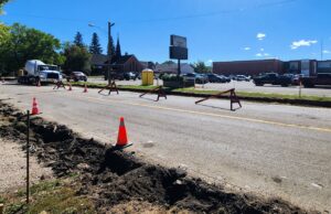 Stettler Back-to-School Traffic: 50 Ave Construction & Detour Map Construction work on a city street with orange cones and red barricades; a deep trench runs along the road and a large cement mixer truck is parked nearby, with buildings and trees in the background.