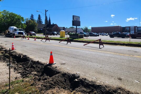 Stettler Back-to-School Traffic: 50 Ave Construction & Detour Map Construction work on a city street with orange cones and red barricades; a deep trench runs along the road and a large cement mixer truck is parked nearby, with buildings and trees in the background.
