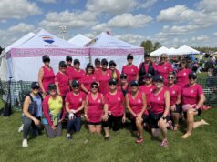 Breasties In The Buff Make Impressive Debut At Edmonton Dragon Boat Festival Group of women in matching pink shirts and hats pose together at an outdoor event under white tents that say 'Breasties in the Buff' on a sunny day.