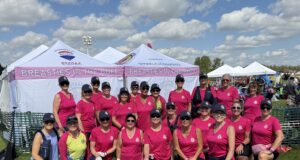 Breasties In The Buff Make Impressive Debut At Edmonton Dragon Boat Festival Group of women in matching pink shirts and hats pose together at an outdoor event under white tents that say 'Breasties in the Buff' on a sunny day.