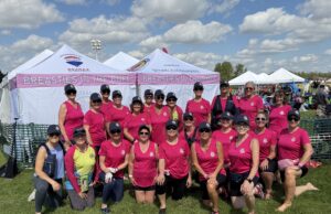 Breasties In The Buff Make Impressive Debut At Edmonton Dragon Boat Festival Group of women in matching pink shirts and hats pose together at an outdoor event under white tents that say 'Breasties in the Buff' on a sunny day.