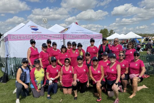 Breasties In The Buff Make Impressive Debut At Edmonton Dragon Boat Festival Group of women in matching pink shirts and hats pose together at an outdoor event under white tents that say 'Breasties in the Buff' on a sunny day.