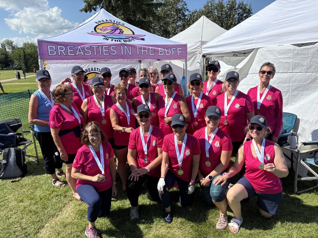 Group of smiling women in pink shirts with medals posing outdoors at a breast cancer charity event under white tents.
