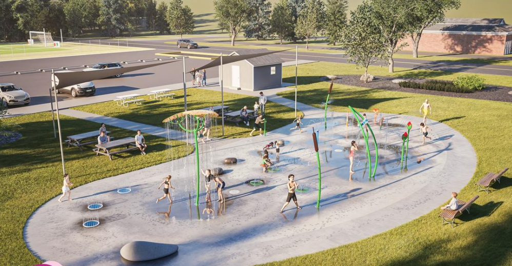 A park splash-pad area where children run through spraying water jets, with benches and a small building in the background.