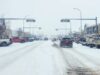 Stettler Town Council Votes To Shorten Downtown Snow Clearing Period Snowy urban street with cars driving and storefronts, snow falling heavily above the road signs.