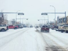 Stettler Town Council Votes To Shorten Downtown Snow Clearing Period Snowy urban street with cars driving and storefronts, snow falling heavily above the road signs.