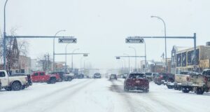 Stettler Town Council Votes To Shorten Downtown Snow Clearing Period Snowy urban street with cars driving and storefronts, snow falling heavily above the road signs.