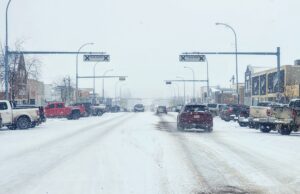 Stettler Town Council Votes To Shorten Downtown Snow Clearing Period Snowy urban street with cars driving and storefronts, snow falling heavily above the road signs.