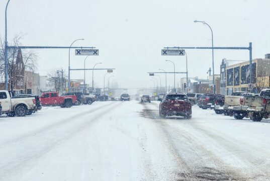 Stettler Town Council Votes To Shorten Downtown Snow Clearing Period Snowy urban street with cars driving and storefronts, snow falling heavily above the road signs.
