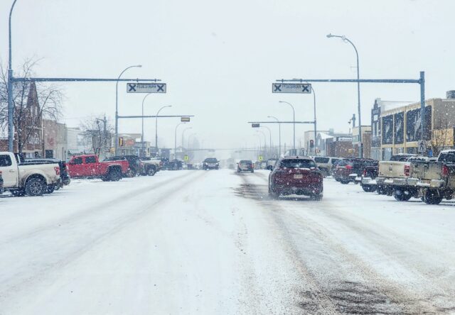 20240321_112319 Snowy urban street with cars driving and storefronts, snow falling heavily above the road signs.