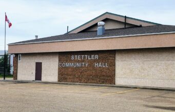 ‘A Seat at the Table’ Seeks Stettler Community Hall Kitchen Access Front view of Stettler Community Hall: beige brick building with 'Stettler Community Hall' sign and a Canadian flag on a flagpole to the left, in an empty parking lot.