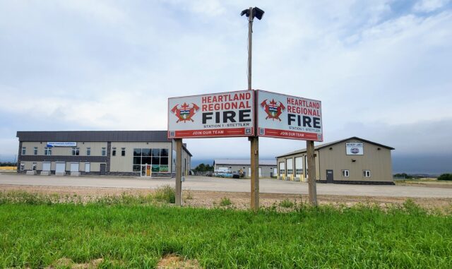 Two Heartland Regional Fire signs mounted on posts in front of a beige commercial building under a cloudy sky, with green grass in the foreground.