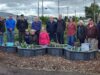 Ceremony Held For Upgraded Stettler Central Community Garden Group of diverse volunteers standing behind raised garden beds in a community garden, with houses and trees in the background.