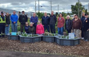 Ceremony Held For Upgraded Stettler Central Community Garden Group of diverse volunteers standing behind raised garden beds in a community garden, with houses and trees in the background.