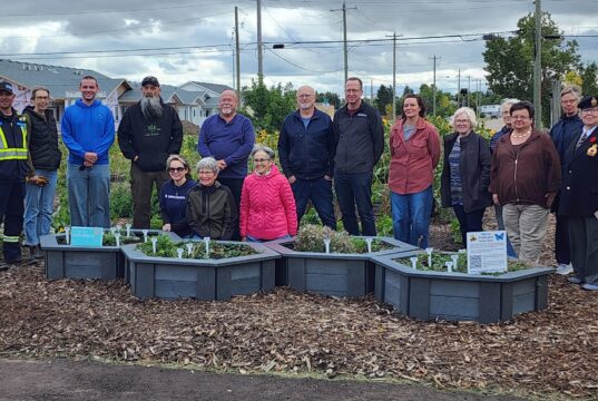 Ceremony Held For Upgraded Stettler Central Community Garden Group of diverse volunteers standing behind raised garden beds in a community garden, with houses and trees in the background.