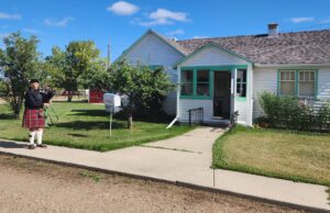 Celebration Of Scottish Culture At The Stettler Town & Country Museum Bagpiper in a kilt and hat playing outside a white house with green trim on a sunny day.