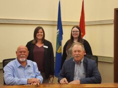 Town and County of Stettler Declare October As Pregnancy & Infant Loss Awareness Month Four adults pose in a conference room, two men seated at a table signing papers while two women stand behind them with flags in the background.