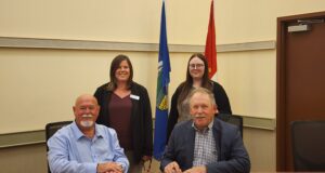 Town and County of Stettler Declare October As Pregnancy & Infant Loss Awareness Month Four adults pose in a conference room, two men seated at a table signing papers while two women stand behind them with flags in the background.