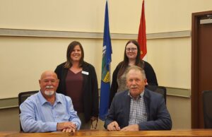 Town and County of Stettler Declare October As Pregnancy & Infant Loss Awareness Month Four adults pose in a conference room, two men seated at a table signing papers while two women stand behind them with flags in the background.