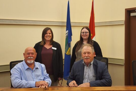 Town and County of Stettler Declare October As Pregnancy & Infant Loss Awareness Month Four adults pose in a conference room, two men seated at a table signing papers while two women stand behind them with flags in the background.