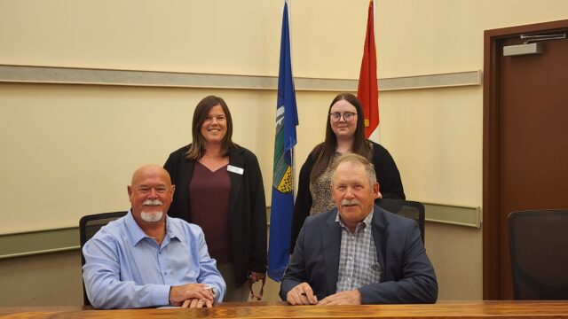 20260921_124238 Four adults pose in a conference room, two men seated at a table signing papers while two women stand behind them with flags in the background.