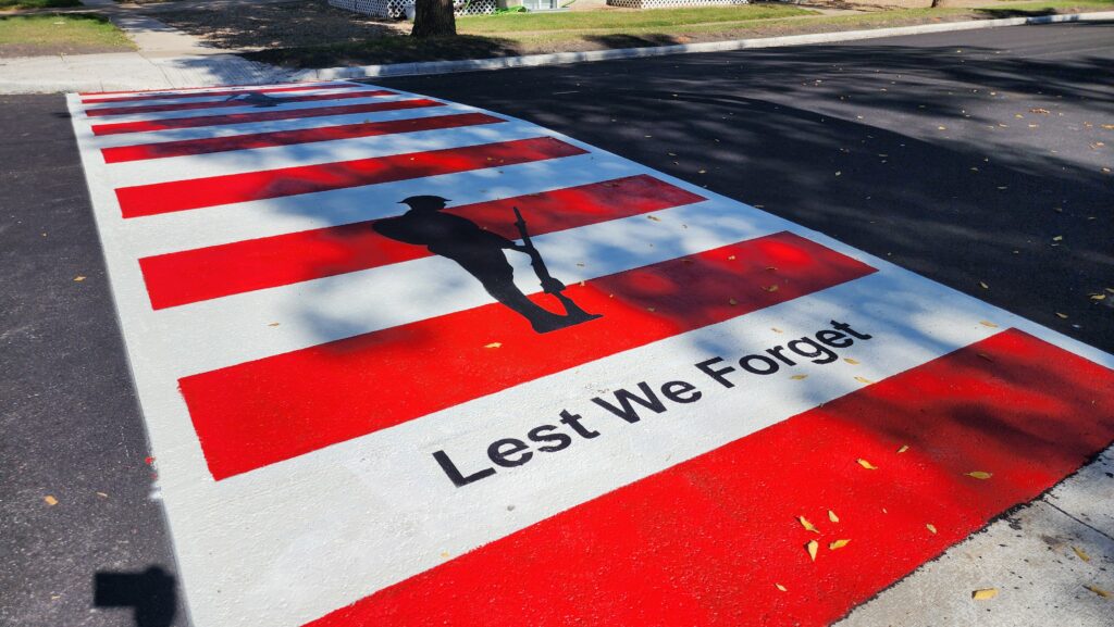 Red-and-white crosswalk mural with a black soldier silhouette and the words "Lest We Forget" across the pavement.