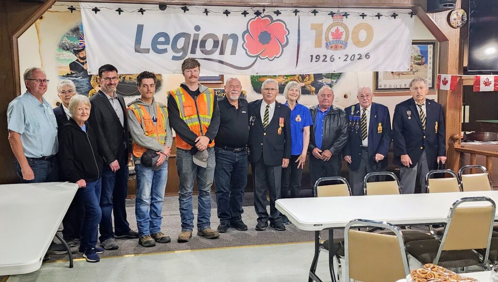 Group of veterans and volunteers posing in front of a Legion banner at a commemorative event, 1926–2026.
