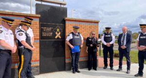 RCMP Unveil Memorial Plaque For Employees Lost To Suicide Group of uniformed officers standing in a semicircle beside a brick memorial wall with a gold-lettered plaque that reads 'Honouring the lives of RCMP employees lost to suicide' and a maple‑leaf ribbon emblem nearby.