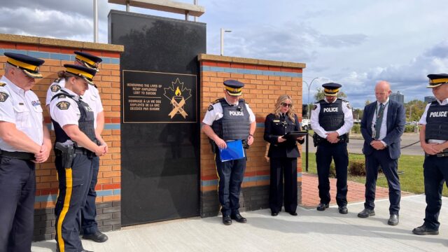 Group of uniformed officers standing in a semicircle beside a brick memorial wall with a gold-lettered plaque that reads 'Honouring the lives of RCMP employees lost to suicide' and a maple‑leaf ribbon emblem nearby.