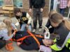 Stettler High School Skills Day Returns October 14 Person lying on a stretcher with orange straps, surrounded by several people outside a brick building, appearing to receive care or assistance.