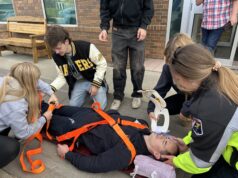 Stettler High School Skills Day Returns October 14 Person lying on a stretcher with orange straps, surrounded by several people outside a brick building, appearing to receive care or assistance.