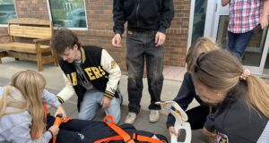 Stettler High School Skills Day Returns October 14 Person lying on a stretcher with orange straps, surrounded by several people outside a brick building, appearing to receive care or assistance.