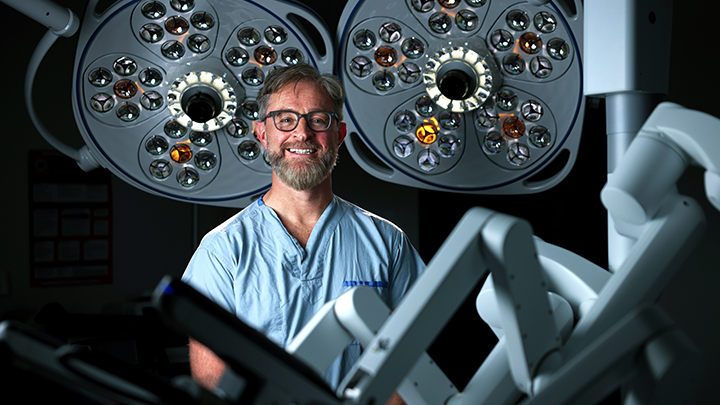 Surgeon in blue scrubs stands under overhead surgical lights in an operating room with robotic equipment near him.