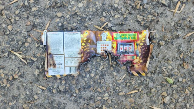 Burnt fireworks wrapper with a Jackpot slot-machine graphic on a rocky, gravel surface.