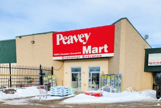 Stettler Home Hardware Moving To Vacant Peavy Mart Location In 2027 Peavey Mart storefront on a beige brick building with a large red sign, snow in the parking lot in front.