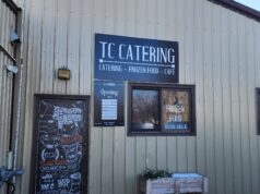 TC Catering Moving To Main Street After Four Years At Auction Mart Exterior of a beige metal building with a large 'TC CATERING' sign and windows displaying menus and chalkboard art outside the shop.