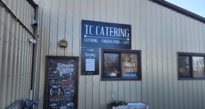 TC Catering Moving To Main Street After Four Years At Auction Mart Exterior of a beige metal building with a large 'TC CATERING' sign and windows displaying menus and chalkboard art outside the shop.