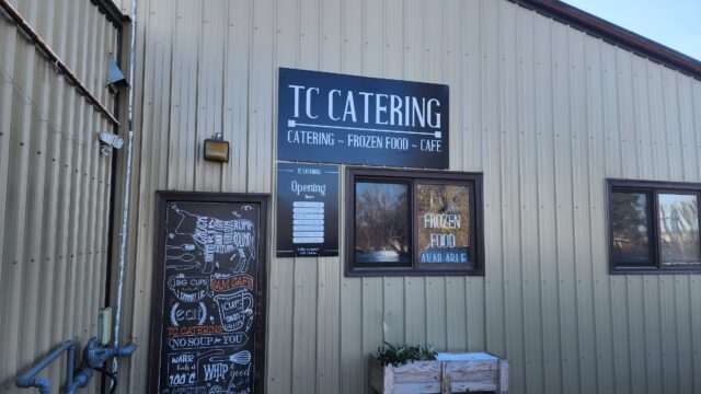 Exterior of a beige metal building with a large 'TC CATERING' sign and windows displaying menus and chalkboard art outside the shop.