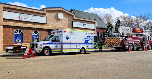 Emergency response scene outside the SALC building with an ambulance and a ladder fire truck as firefighters work on site nearby.