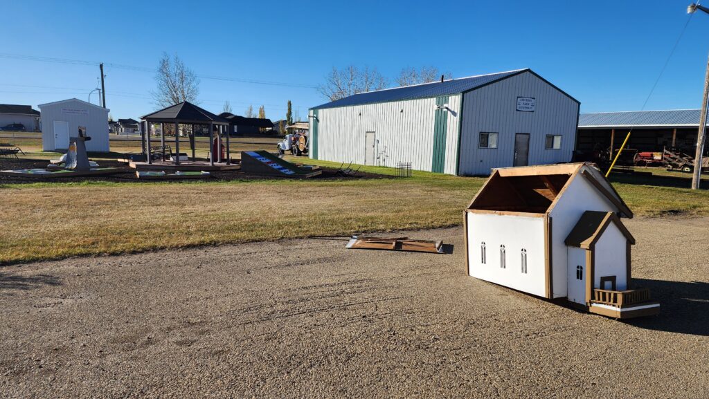 Outdoor display of scale-model buildings, including white houses and a pavilion, set on a grassy field at a rural yard.
