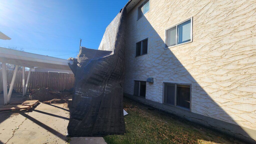 Two-story beige stucco building with a large, leaning wooden beam and scattered construction debris in the yard under a clear blue sky